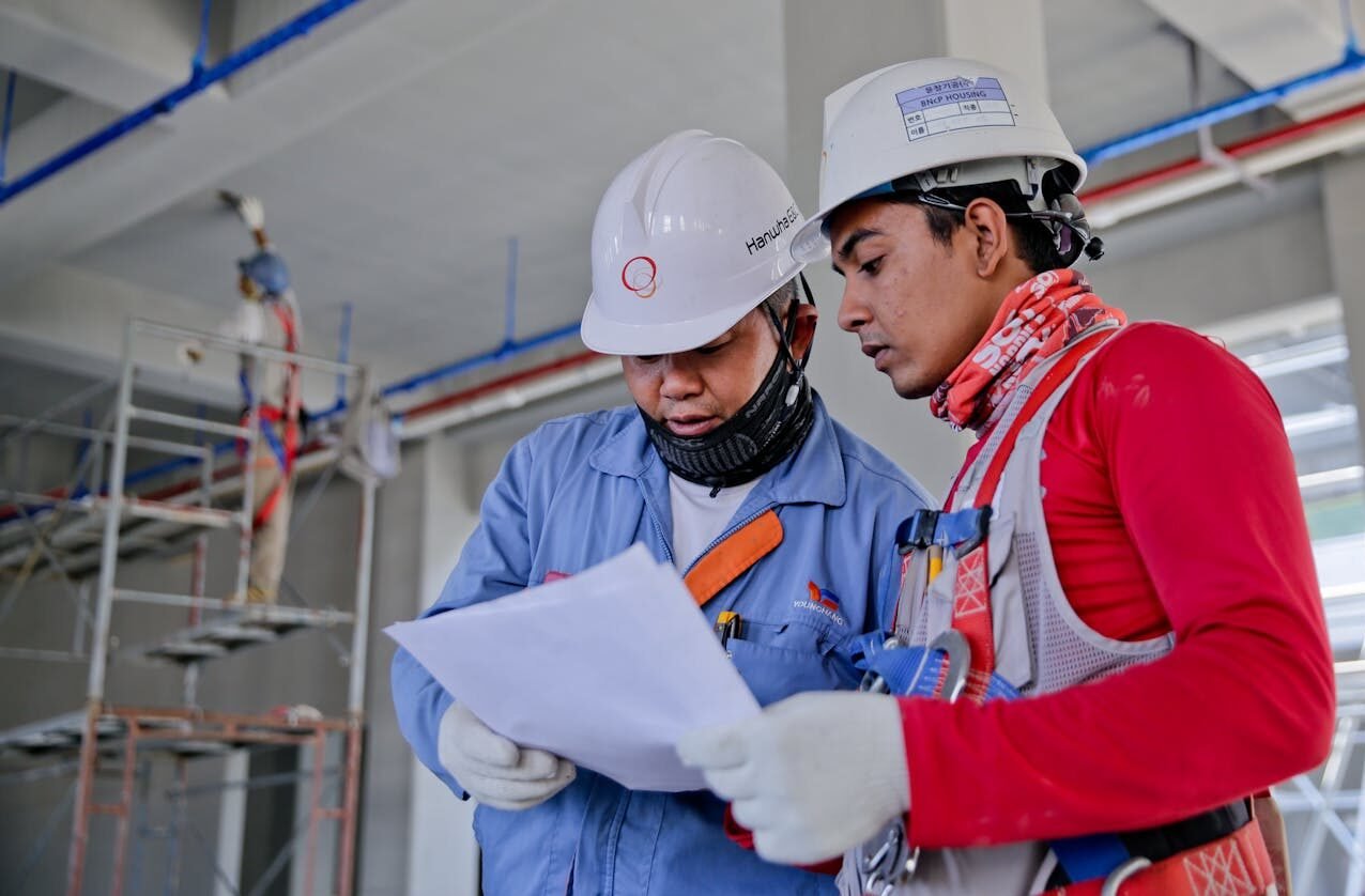Two engineers in safety helmets reviewing construction plans at a worksite.